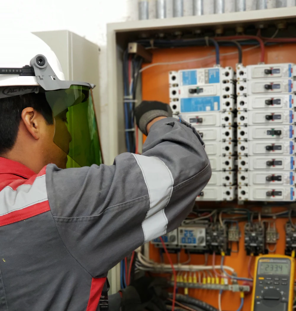 An electrician in a hard hat and face shield testing a panel